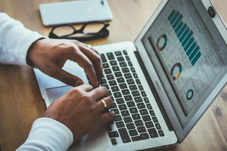Nell'immagine, il primo piano sulle mani di un uomo intento a lavorare ad un laptop. Alla sua sinistra, ci sono un paio di occhiali e un cellulare poggiati sul tavolo.